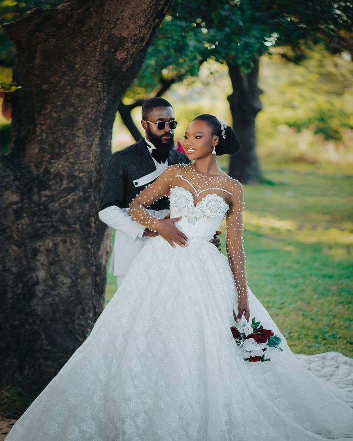 gallery-06 Bride and groom pose under a tree in elegant wedding attire outdoors.